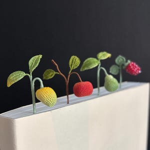 Puede incluir: Conjunto de marcadores de ganchillo hechos a mano con frutas y hojas. Los marcadores incluyen un limón amarillo, una manzana roja, una pera verde y una frambuesa roja. Cada marcador está unido a un tallo y hojas.