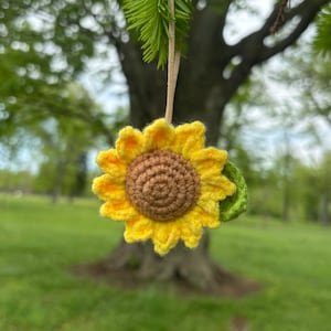以下が含まれることがあります： 黄色の花びら、茶色の中心、緑の葉が付いた、かぎ針編みのヒマワリのオーナメント。 手作りの装飾は、自然色の紐から吊り下げられており、あらゆる空間に自然のタッチを加えるのに最適です。