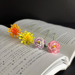 May include: Four crocheted flowers in different colors, including orange, yellow, purple, and pink, are arranged on an open book. The flowers are delicate and intricate, with detailed stitching.