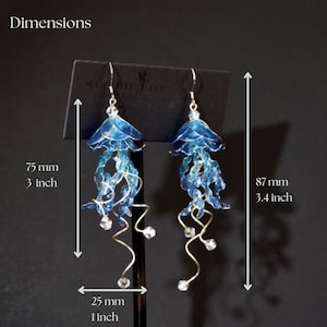 以下が含まれることがあります： シルバーのフックとアクセントが付いた、青いクラゲのイヤリングのペア。イヤリングの長さは7.5cm、幅は2.5cmで、透明なクリスタルのようなビーズが付いています。暗い背景に表示されています。