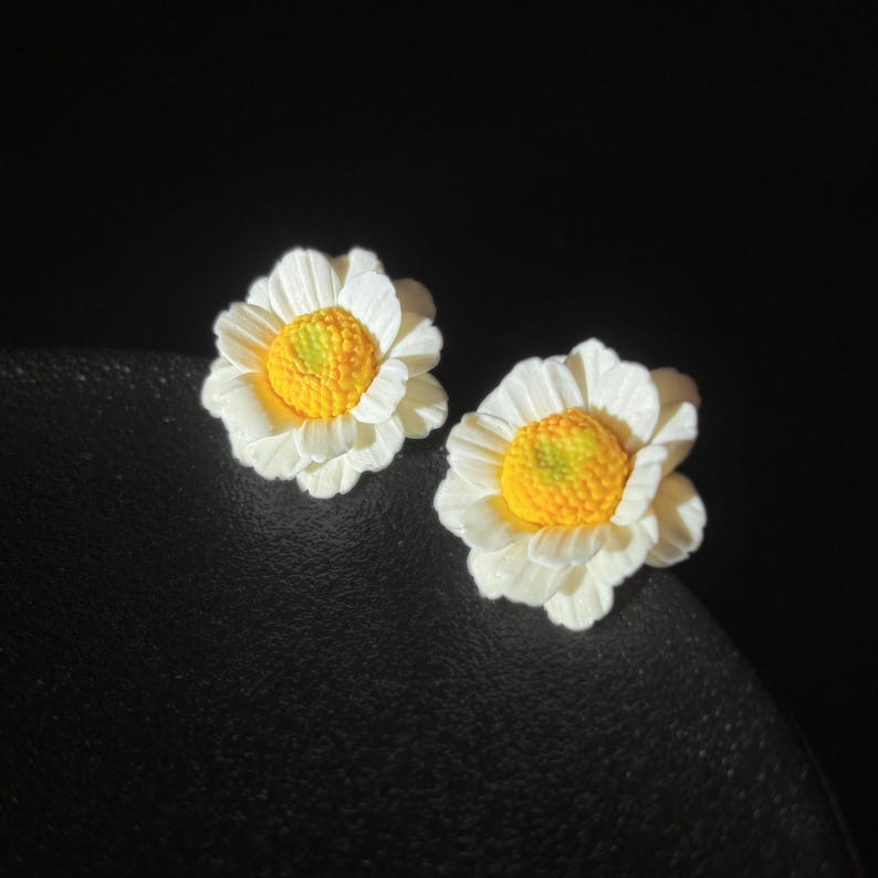 以下が含まれることがあります： 白いデイジーの花の形をしたスタッドピアス一対。質感のある花びらと、盛り上がった鮮やかな黄色の中心部が特徴です。花のデザインは重なり合った花びらで立体感を出し、細部までこだわったリアルな植物の美しさを表現しています。