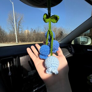 May include: Crocheted car charm featuring three blue flower-shaped ornaments. The charm is suspended from a green stem and bow, adding a touch of handmade charm to any vehicle's interior. The flowers are in varying shades of blue.