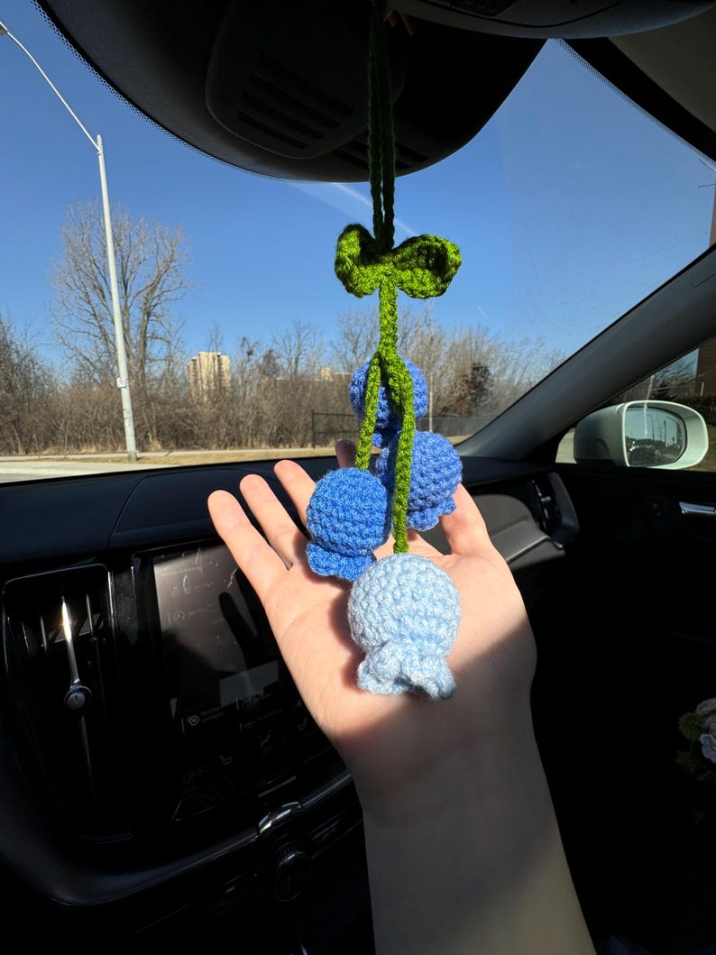 May include: Crocheted car charm featuring three blue flower-shaped ornaments. The charm is suspended from a green stem and bow, adding a touch of handmade charm to any vehicle's interior. The flowers are in varying shades of blue.