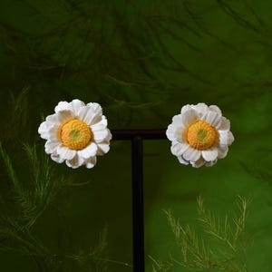 以下が含まれることがあります： 質感のある黄色い中心部を持つ白いデイジーの花の形をしたスタッドピアス一対。各ピアスは、詳細で丸みを帯びた黄色い芯を囲む層状の白い花びらが特徴で、暗い垂直のディスプレイスタンドに取り付けられています。