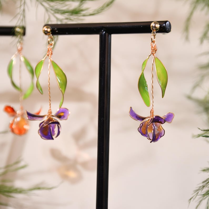 May include: Dangle earrings featuring a flower design with translucent purple and orange petals, accented by bright green leaves. The floral elements are crafted from thin wire and suspended from gold-toned metal posts on a black display stand.