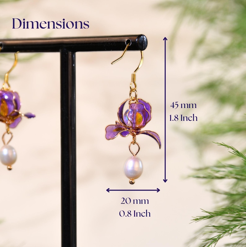 May include: A pair of gold-toned hook earrings featuring purple translucent flower petals and a dangling white pearl. The text Dimensions is visible, with measurements of 1.8 inches in height and 0.8 inches in width indicated by arrows.