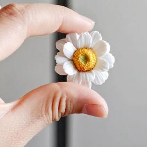 以下が含まれることがあります： 複数の白い丸みを帯びた花びらが、質感のある黄金色の中心部の周りに円形に配置された、小さな3次元のデイジーの花のオーナメント。この花は、深みのある層状でリアルな外観をしています。
