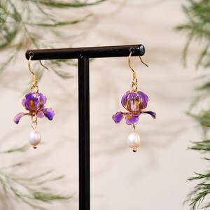 May include: A pair of drop earrings featuring translucent purple flower-shaped charms with gold-toned wire edges. Each earring is suspended from a gold-toned hook and finished with a single white, oval-shaped pearl dangling from the base of the flower.