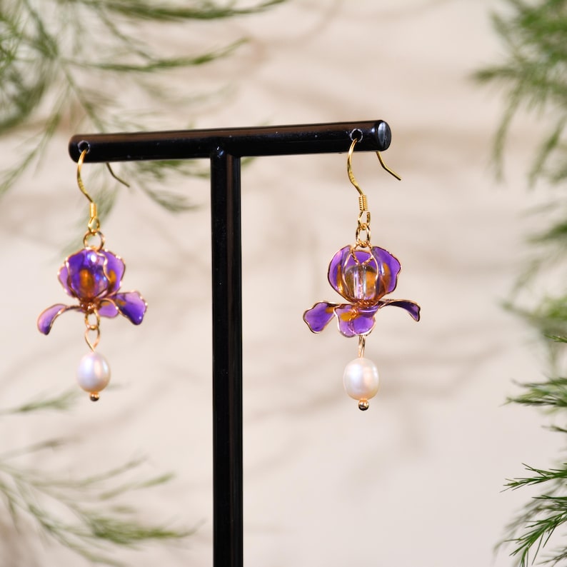 May include: A pair of drop earrings featuring translucent purple flower-shaped charms with gold-toned wire edges. Each earring is suspended from a gold-toned hook and finished with a single white, oval-shaped pearl dangling from the base of the flower.