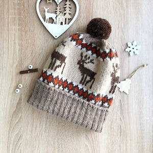 Può includere: Un cappello di lana marrone con un pon pon in cima. Il cappello ha un motivo bianco e arancione con renne e fiocchi di neve.