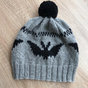 Può includere: Un cappello di lana grigio con un pon pon nero in cima. Il cappello ha un disegno di pipistrello nero sul davanti.