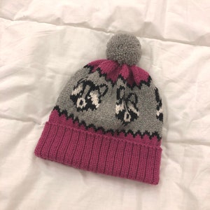 Puede incluir: Un gorro de punto gris y rosa con un pomp&oacute;n en la parte superior. El gorro tiene un patr&oacute;n de mapaches.