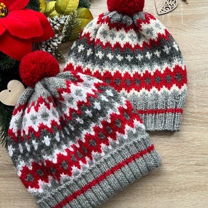 Può includere: Due berretti invernali lavorati a maglia con pompon rossi. I berretti presentano un motivo geometrico grigio, bianco e rosso. I berretti sono realizzati in un materiale morbido e caldo, ideale per il freddo. I cappelli sono esposti su una superficie di legno.