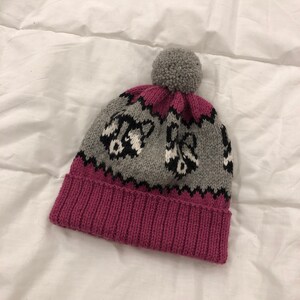 Puede incluir: Un gorro de punto gris y rosa con un pomp&oacute;n en la parte superior. El gorro tiene un patr&oacute;n de mapaches negros y blancos en la parte gris.
