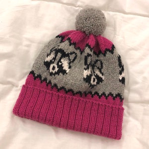 Puede incluir: Un gorro de punto rosa y gris con un pomp&oacute;n en la parte superior. El gorro tiene un patr&oacute;n de mapaches en blanco y negro.