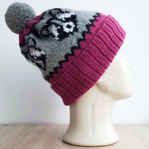 Puede incluir: Un gorro de punto rosa y gris con un pomp&oacute;n blanco en la parte superior. El gorro tiene un patr&oacute;n geom&eacute;trico y est&aacute; hecho de lana.