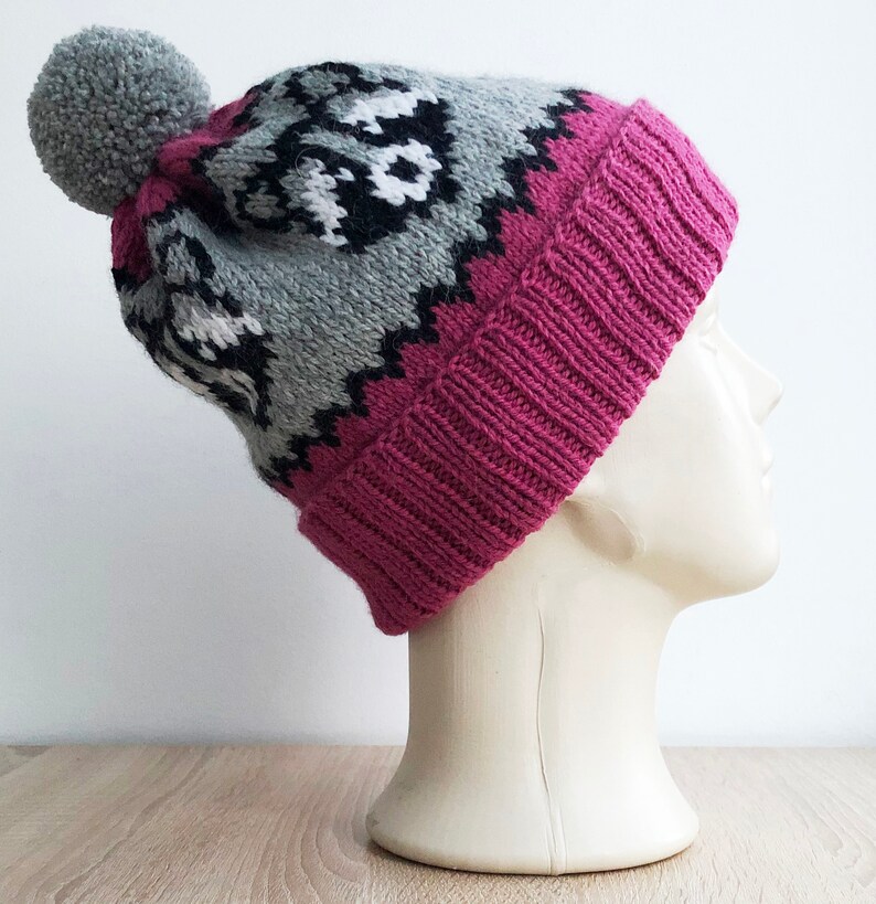 Puede incluir: Un gorro de punto rosa y gris con un pomp&oacute;n blanco en la parte superior. El gorro tiene un patr&oacute;n geom&eacute;trico y est&aacute; hecho de lana.