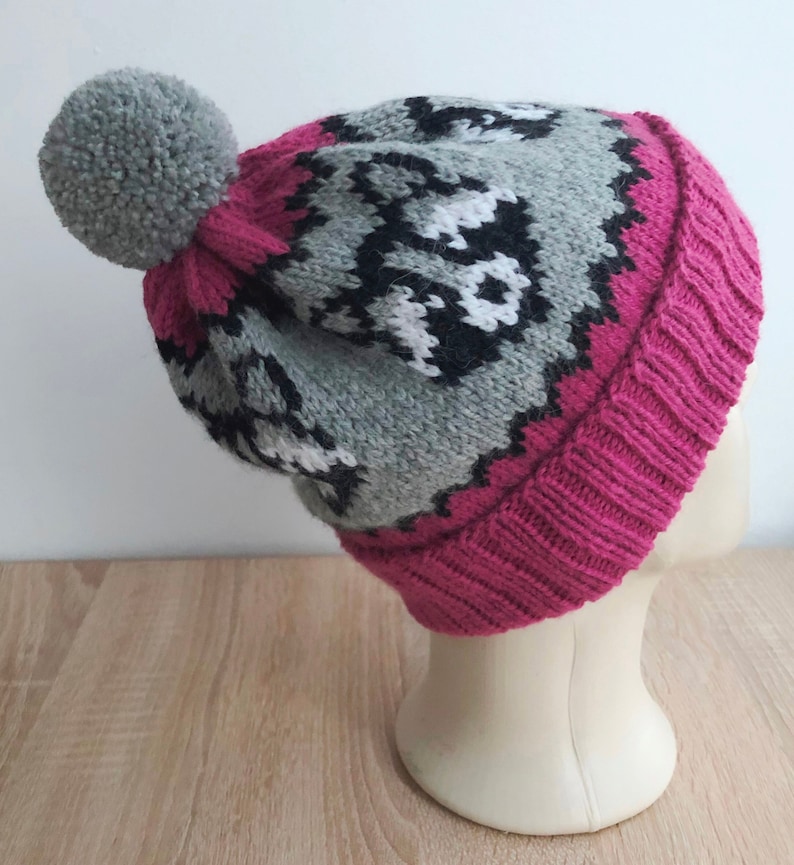 Puede incluir: Un gorro de punto gris y rosa con un patr&oacute;n geom&eacute;trico blanco y negro. El gorro tiene un pomp&oacute;n gris esponjoso en la parte superior.