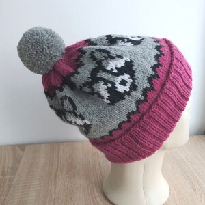 Puede incluir: Un gorro de punto gris y rosa con un pomp&oacute;n blanco en la parte superior. El gorro tiene un patr&oacute;n geom&eacute;trico con detalles en blanco y negro.