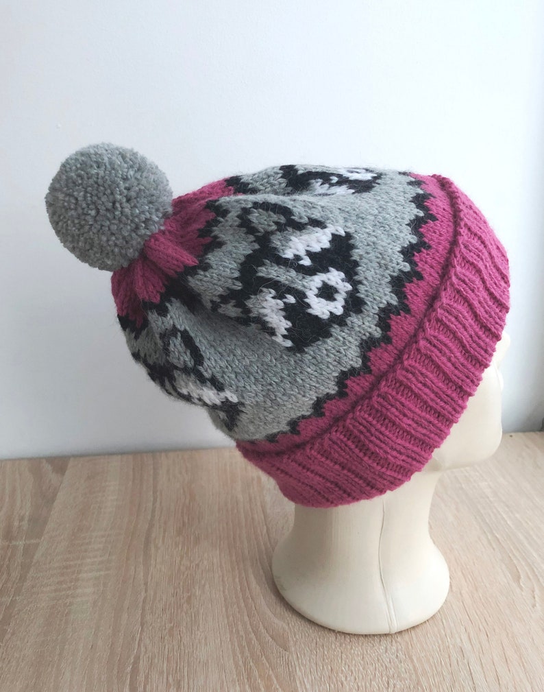 Puede incluir: Un gorro de punto gris y rosa con un pomp&oacute;n blanco en la parte superior. El gorro tiene un patr&oacute;n geom&eacute;trico con detalles en blanco y negro.