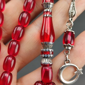 May include: A red prayer beads necklace with silver accents. The beads are oval shaped and the pendant is a crescent moon with a star.