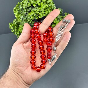 May include: A red amber prayer beads necklace with silver accents. The beads are round and have a glossy finish. The necklace is strung on a silver chain with a silver tassel.