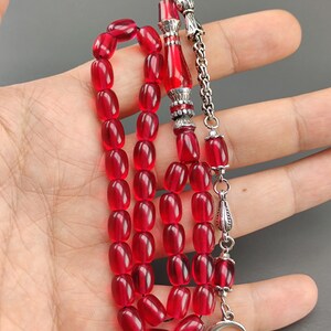 May include: A red prayer bead necklace with silver accents. The beads are oval shaped and the necklace has a silver tassel.