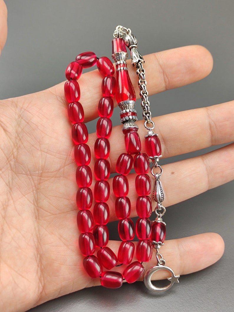 May include: A red prayer bead necklace with silver accents. The beads are oval shaped and the necklace has a silver tassel.