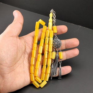 Op de afbeelding: Een gele gebedsketting met een zilveren kwast en een decoratieve hanger. De kralen zijn rechthoekig en doorschijnend. De kwast en hanger zijn zilver. De kralen worden in een hand gehouden tegen een zwarte achtergrond.