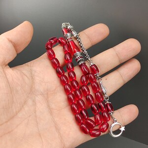 May include: A red prayer beads necklace with silver accents. The beads are oval shaped and the necklace has a silver tassel with a crescent moon and star charm.
