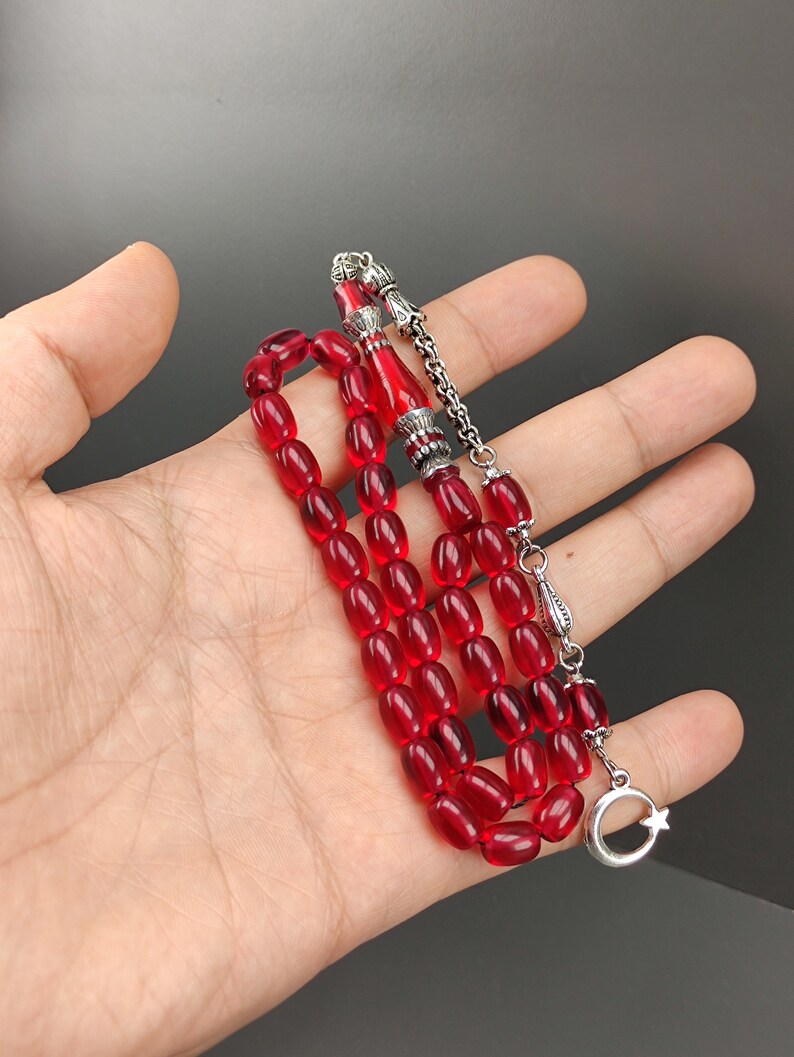 May include: A red prayer beads necklace with silver accents. The beads are oval shaped and the necklace has a silver tassel with a crescent moon and star charm.