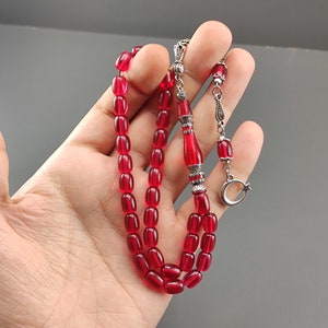 May include: A red prayer bead necklace with silver accents. The beads are oval shaped and the necklace has a silver tassel and a crescent moon charm.