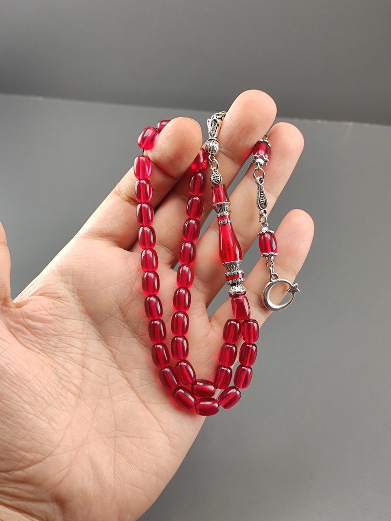 May include: A red prayer bead necklace with silver accents. The beads are oval shaped and the necklace has a silver tassel and a crescent moon charm.