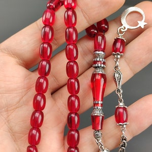 May include: A red prayer beads necklace with silver accents. The beads are oval shaped and the necklace has a silver tassel and a silver charm with a crescent moon and star.