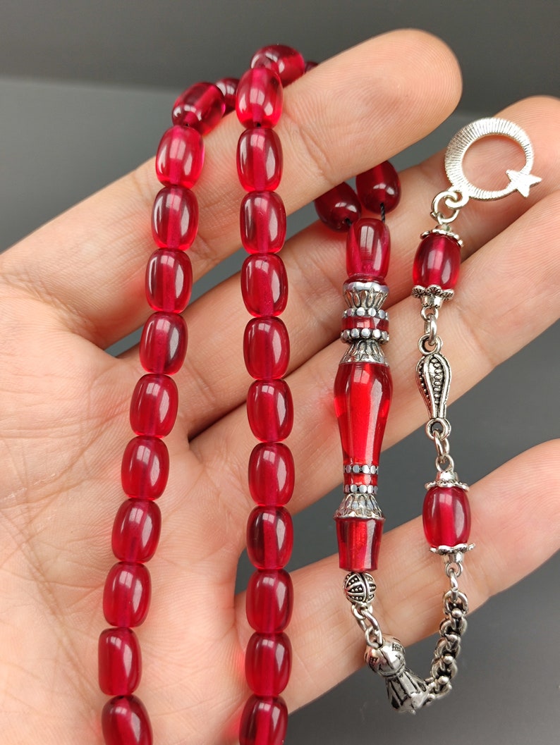 May include: A red prayer beads necklace with silver accents. The beads are oval shaped and the necklace has a silver tassel and a silver charm with a crescent moon and star.