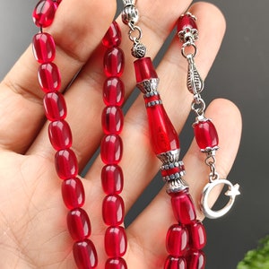 May include: A red prayer beads necklace with silver accents. The beads are oval shaped and have a glossy finish. The necklace has a silver tassel and a crescent moon charm.