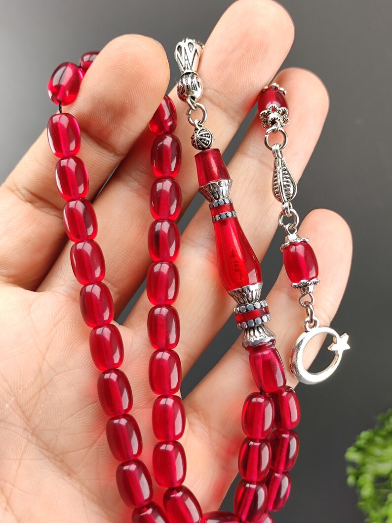 May include: A red prayer beads necklace with silver accents. The beads are oval shaped and have a glossy finish. The necklace has a silver tassel and a crescent moon charm.