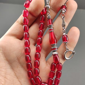 May include: A red prayer bead necklace with silver accents. The beads are oval shaped and the necklace has a silver tassel with a crescent moon charm.