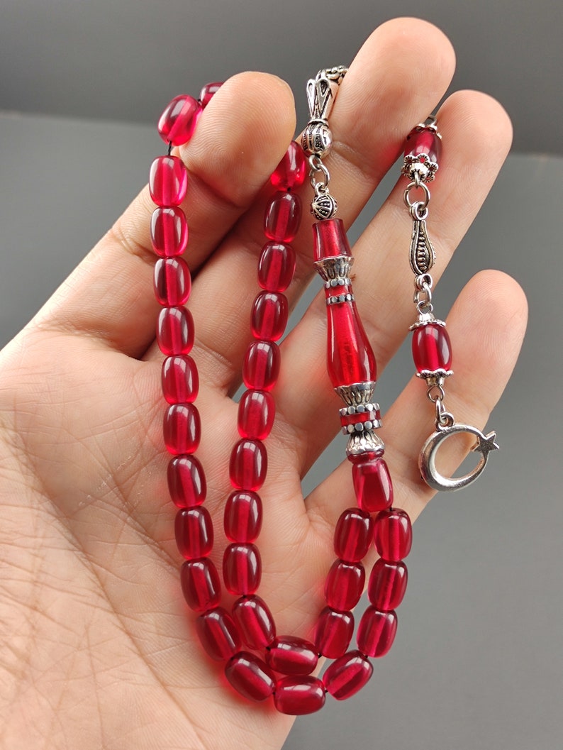 May include: A red prayer bead necklace with silver accents. The beads are oval shaped and the necklace has a silver tassel with a crescent moon charm.