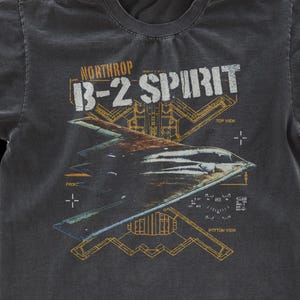 May include: Dark gray t-shirt featuring a graphic of a B-2 Spirit stealth bomber with the text "NORTHROP B-2 SPIRIT". The design includes schematics and views of the aircraft in yellow and white.