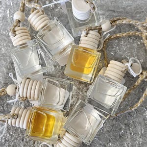 May include: A collection of small, square glass bottles with wooden tops and decorative cords. Some bottles contain a clear liquid, while others have a golden-colored liquid. The bottles are arranged on a textured, reflective surface.