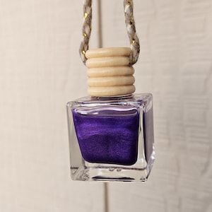 Puede incluir: Botella cuadrada de vidrio transparente con tapón de madera y cuerda trenzada para colgar. Contiene un líquido morado oscuro y brillante. La cuerda es gris con detalles dorados. Fondo claro.