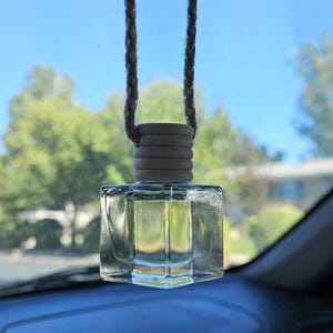 Könnte beinhalten: Transparenter, quadratischer Auto-Lufterfrischer, der an einer Schnur hängt. Der Lufterfrischer hat eine hellfarbige Oberseite und eine transparente Glasflasche. Der Hintergrund zeigt einen blauen Himmel und grüne Bäume.