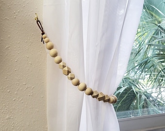 May include: A wooden beaded curtain tieback in a light brown color. The tieback is looped around a white sheer curtain.