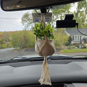 May include: A small, white ceramic pot with a green plant hangs from a macrame plant hanger. The plant is suspended from the rearview mirror inside a vehicle. The macrame is beige and has a tassel at the bottom.