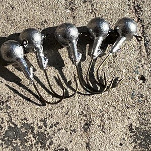 May include: Five silver-colored fishing jig heads with attached bronze-colored hooks are arranged on a textured, gray surface. The jig heads have a rounded shape with a pointed hook attachment.