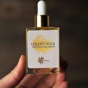May include: A clear glass bottle of Golden Hour Soothing Face Serum. The bottle has a white label with gold text and a watercolor-style yellow design. The dropper is white and gold. The product is held in a hand.