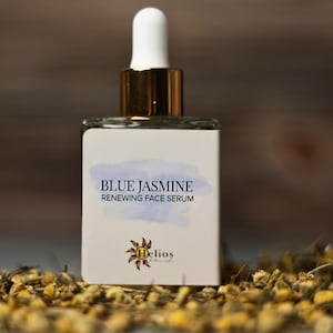 Op de afbeelding: Een vierkante glazen fles Blue Jasmine Renewing Face Serum met een witte druppelkop en gouden accenten. Het etiket toont de productnaam en een logo. De fles is omgeven door gedroogde bloemen.