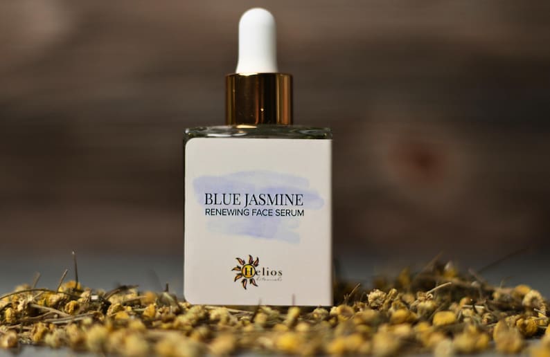 Op de afbeelding: Een vierkante glazen fles Blue Jasmine Renewing Face Serum met een witte druppelkop en gouden accenten. Het etiket toont de productnaam en een logo. De fles is omgeven door gedroogde bloemen.