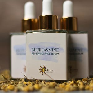 Op de afbeelding: Drie glazen flessen Blue Jasmine Renewing Face Serum met witte druppelaars en gouden accenten. De etiketten tonen de productnaam en een aquarelontwerp. De flessen zijn omgeven door gedroogde bloemen.